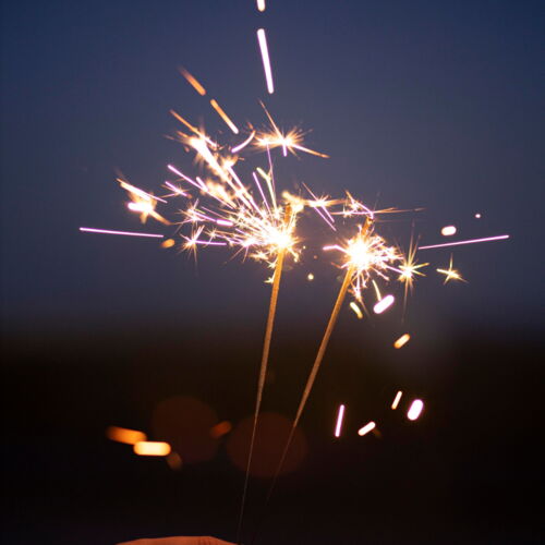 Eine Hand halt zwei brennende Wunderkerzen vor einem abendlich dunkelblauen Himmel.